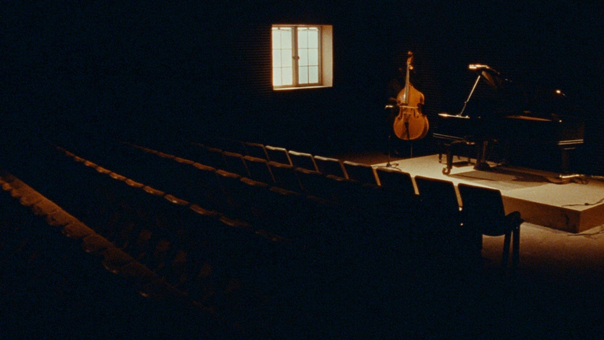 An intimate empty jazz listening room before doors open, seats angled toward a small low stage under a single warm light.