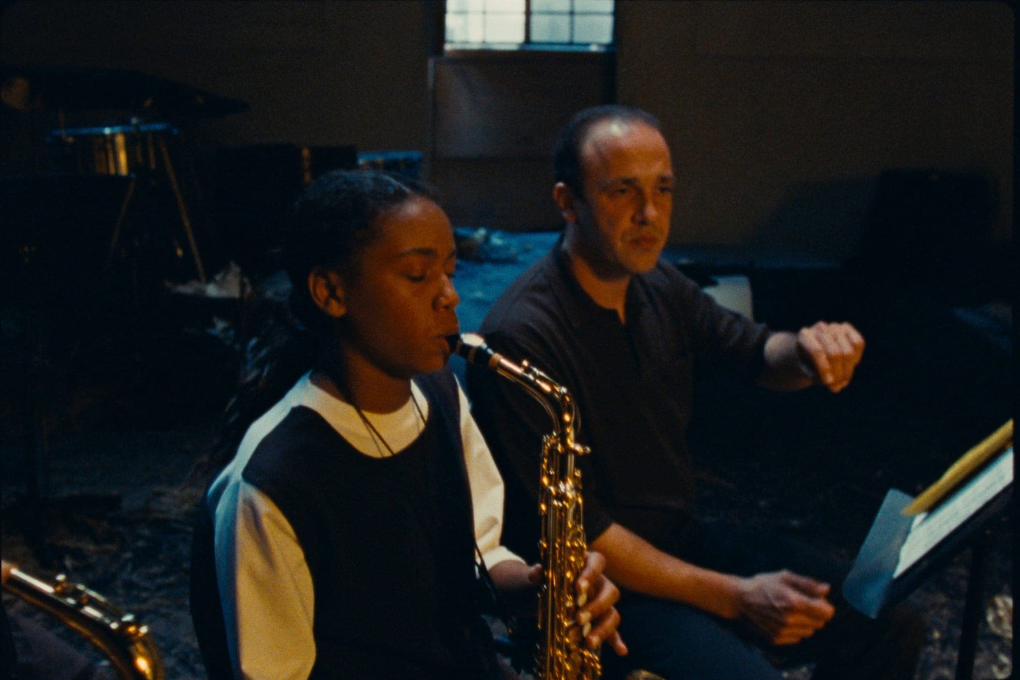 A teenage saxophonist mid-phrase in a rehearsal room, an adult mentor listening beside them.